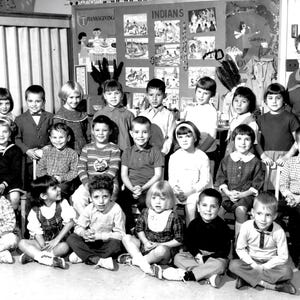 May include: Black and white photograph of a group of children and a teacher in a classroom. The children are seated and standing, wearing various outfits. A sign on the wall reads "Indians".