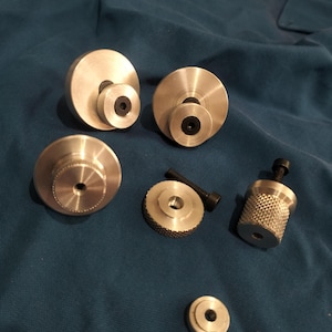 Può includere: Una collezione di componenti metallici color argento che include dischi circolari con fori centrali, alcuni con bordi zigrinati, e manopole cilindriche con lati testurizzati. Diversi pezzi includono viti in metallo nero inserite al centro.