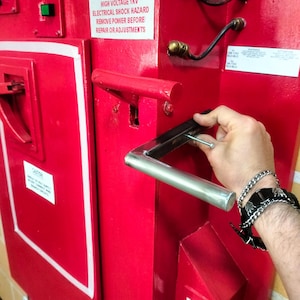 Può includere: Un pannello elettrico rosso con una maniglia argentata e il testo "DANGER HIGH VOLTAGE 1KV ELECTRICAL SHOCK HAZARD REMOVE POWER BEFORE REPAIR OR ADJUSTMENTS". Una mano tiene una vite vicino alla maniglia.