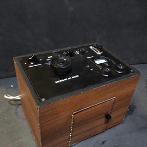 May include: A vintage Beckman pH meter in a wooden cabinet with a black control panel. The panel has dials, knobs, and a small display. The text "BECKMAN pH METER" is visible. A power cord extends from the side of the unit.