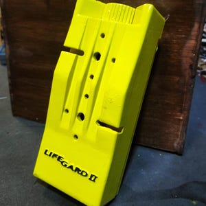 May include: A bright yellow plastic box with the words "LIFEGUARD II" printed in black. The box has a textured surface and several small holes.