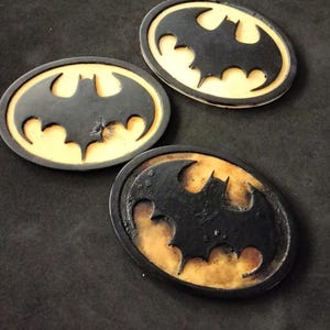 Op de afbeelding: Drie ovale Batman-logo-emblemen. Elk embleem heeft een zwart vleermuissilhouet in een gele ovale rand. De emblemen hebben een getextureerd, mogelijk leerachtig uiterlijk, met verschillende gradaties van slijtage.