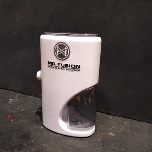 May include: A white "Mr. Fusion Home Energy Reactor" with a clear, curved front section. The device has a circular logo with crossed bones and text. A small knob is on the side.