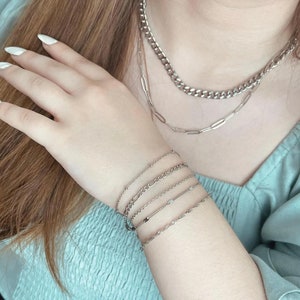 May include: A close-up of a person's wrist wearing five silver chain bracelets. The bracelets are layered and have different chain styles.