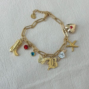 Puede incluir: Pulsera de oro con una variedad de dijes, que incluyen una bota, una mariposa, un ojo, un corazón, una estrella y una letra B.