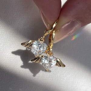 A close-up of a hand holding a pair of gold angel wings heart earrings, featuring sparkling cubic zirconia heart charms. The delicate wing details and radiant stones catch the sunlight, casting a subtle shadow on the white surface. These hypoallergenic earrings are perfect for everyday wear or as a meaningful gift for Christmas, birthdays, or special occasions. A beautiful choice for those who love celestial and angel-inspired jewelry.