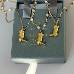 Pode incluir: Três colares de ouro com pingentes de botas em uma caixa cinza com o texto "Rachel Rose" e "Estd 2022!" Os colares são adornados com contas de pérolas.