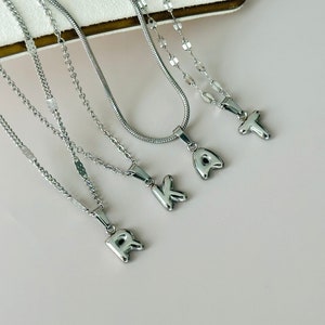 May include: Four silver-toned metal necklaces, each featuring a single letter pendant. The pendants are shaped like the letters R, K, A, and T. The necklaces consist of various chain styles, including a snake chain and delicate link chains.