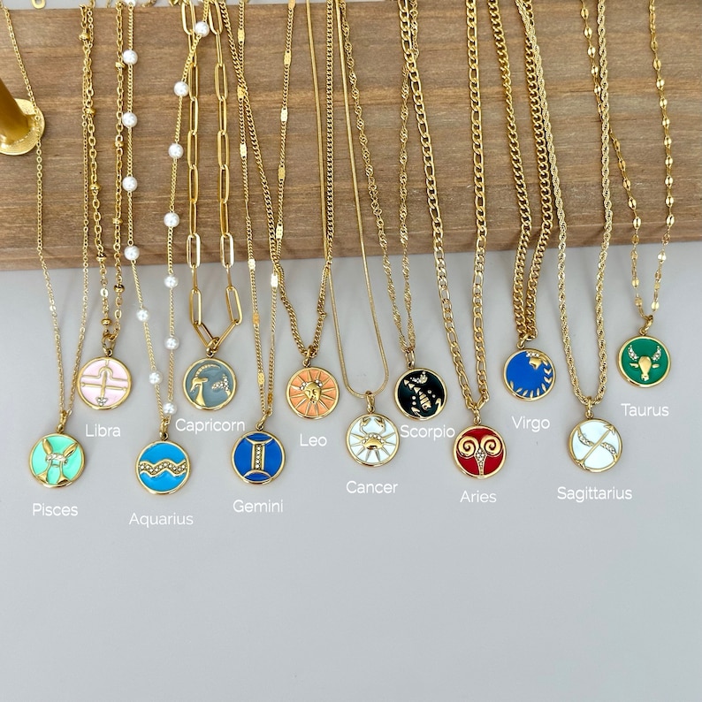 K&ouml;nnte beinhalten: Goldfarbene Halsketten mit verschiedenen Sternzeichen-Anh&auml;ngern. Jeder Anh&auml;nger hat ein farbenfrohes Emaille-Design und ist mit seinem entsprechenden Sternzeichen beschriftet. Die Halsketten werden vor einem Holzgrund pr&auml;sentiert.