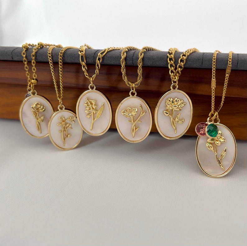 May include: Six gold-toned necklaces featuring oval pendants with white mother-of-pearl backgrounds and raised gold floral designs. The rightmost pendant includes two small round crystals in pink and green attached to the top of the oval frame.