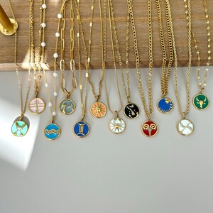 K&ouml;nnte beinhalten: Eine Sammlung goldfarbener Halsketten mit verschiedenen Anh&auml;ngerdesigns, darunter Tierkreiszeichen und geometrische Formen. Die Anh&auml;nger sind mit Emaille in Farben wie Blau, Rosa und Gr&uuml;n versehen. Die Halsketten werden auf einer Holzoberfl&auml;che pr&auml;sentiert.