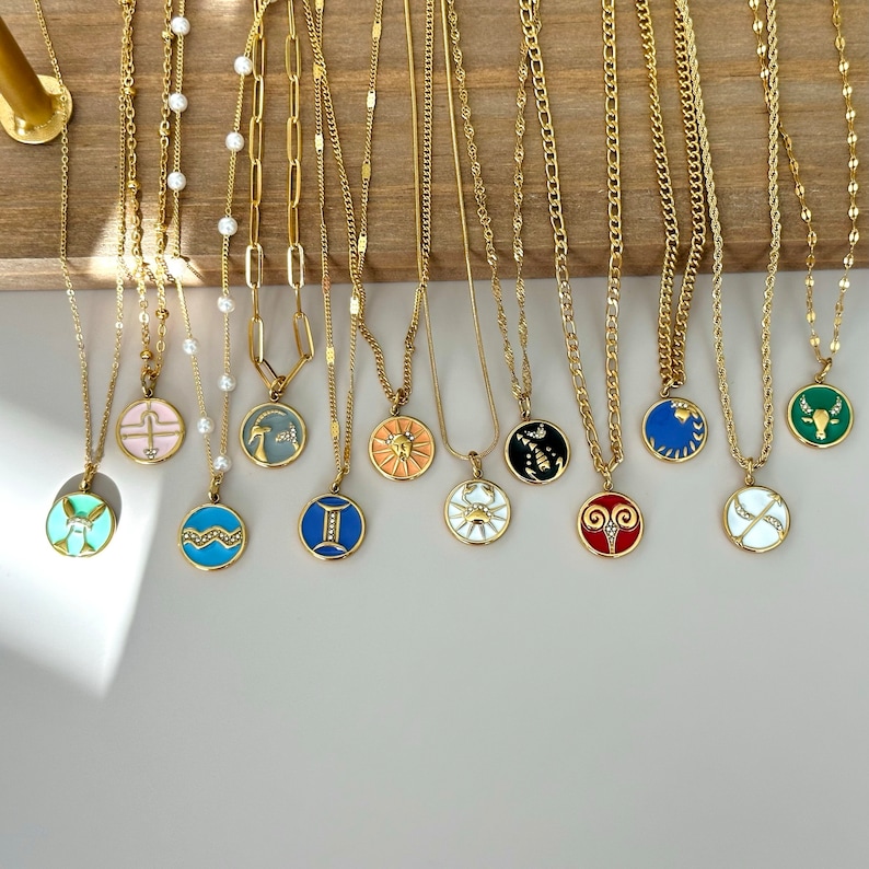 K&ouml;nnte beinhalten: Eine Sammlung goldfarbener Halsketten mit verschiedenen Anh&auml;ngerdesigns, darunter Tierkreiszeichen und geometrische Formen. Die Anh&auml;nger sind mit Emaille in Farben wie Blau, Rosa und Gr&uuml;n versehen. Die Halsketten werden auf einer Holzoberfl&auml;che pr&auml;sentiert.