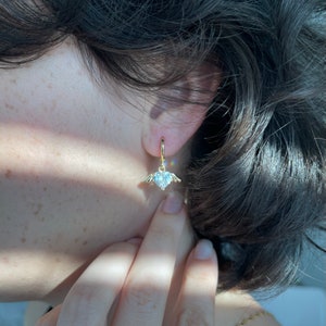 Może przedstawiać: Złote kolczyki koła z wiszącym sercem ozdobionym przezroczystym kryształem i małymi skrzydłami.