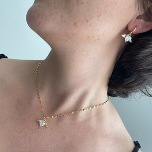 Może przedstawiać: Złoty łańcuszek z małym serduszkowym wisiorkiem ozdobionym przezroczystym kryształem i złotymi skrzydłami. Pasujące złote kolczyki z przezroczystym kryształem i złotymi skrzydłami.