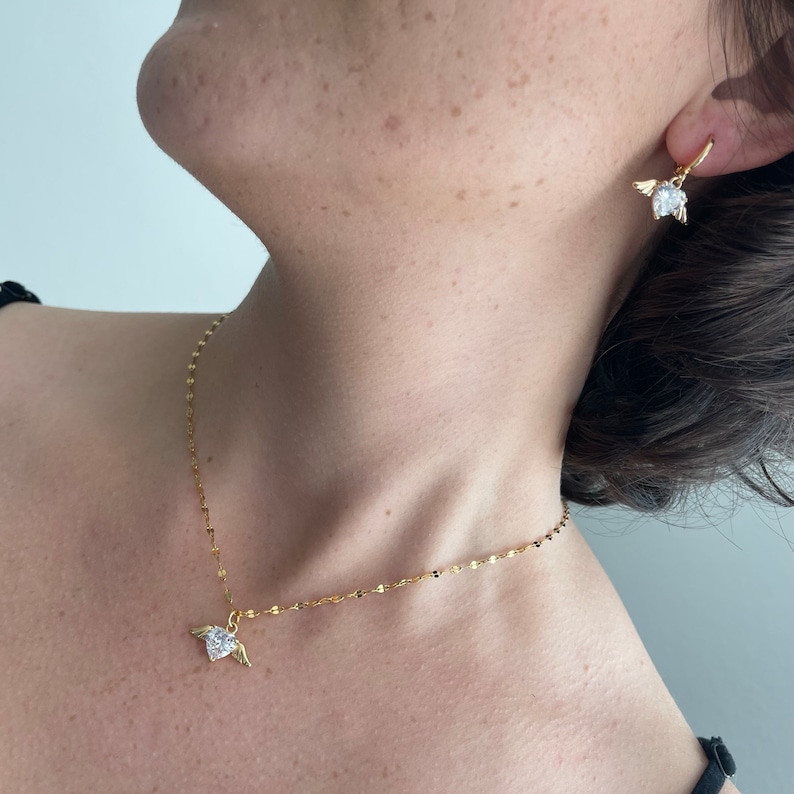 Może przedstawiać: Złoty łańcuszek z małym serduszkowym wisiorkiem ozdobionym przezroczystym kryształem i złotymi skrzydłami. Pasujące złote kolczyki z przezroczystym kryształem i złotymi skrzydłami.