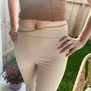 May include: A gold chain belt with the text "6mm Curb" on it. The belt is worn around the waist of a person wearing beige pants.