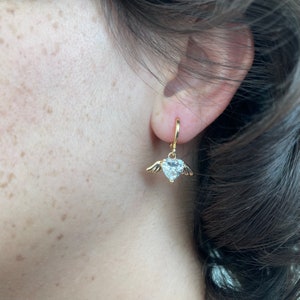 Może przedstawiać: Złote kolczyki koła z wiszącym sercem z kryształu ozdobionym małymi skrzydłami.