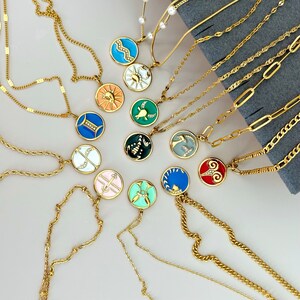 K&ouml;nnte beinhalten: Eine Sammlung goldfarbener Halsketten mit verschiedenen Anh&auml;ngerdesigns. Die runden Anh&auml;nger sind mit farbenfrohen Emaille-Designs versehen, darunter Tierkreiszeichen. Die Halsketten werden auf einer hellen Oberfl&auml;che pr&auml;sentiert.