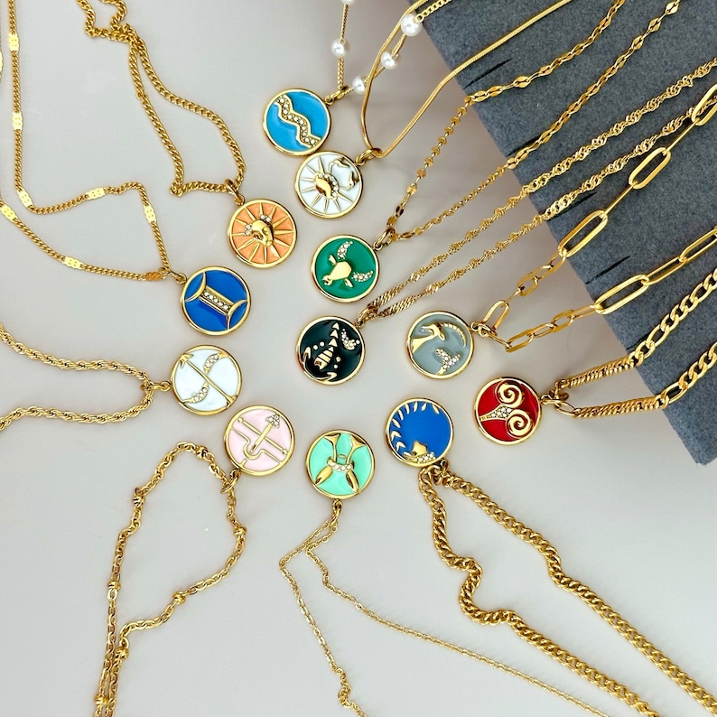 K&ouml;nnte beinhalten: Eine Sammlung goldfarbener Halsketten mit verschiedenen Anh&auml;ngerdesigns. Die runden Anh&auml;nger sind mit farbenfrohen Emaille-Designs versehen, darunter Tierkreiszeichen. Die Halsketten werden auf einer hellen Oberfl&auml;che pr&auml;sentiert.