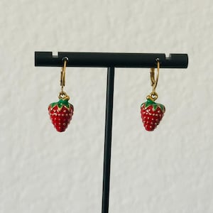 May include: A pair of strawberry-shaped earrings. Each earring features a red, textured strawberry with green leaves and gold-colored accents. The earrings are suspended from gold-colored hoops.