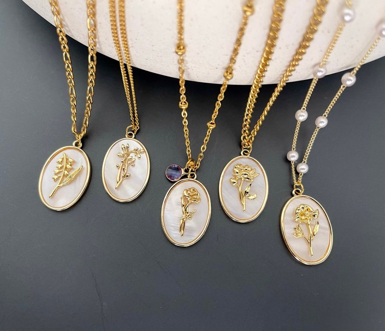 May include: Five gold-toned necklaces featuring oval pendants with white iridescent backgrounds and raised gold floral motifs. The chains vary in style, including link chains and one chain accented with small white pearls and a small purple crystal charm.
