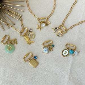 May include: Gold-toned necklaces and charms with various designs. Charms include a jade pendant, a cowboy boot, a coin, a square with a blue gem, a letter "R" pendant, and a starfish. The necklaces are on a cream-colored surface.