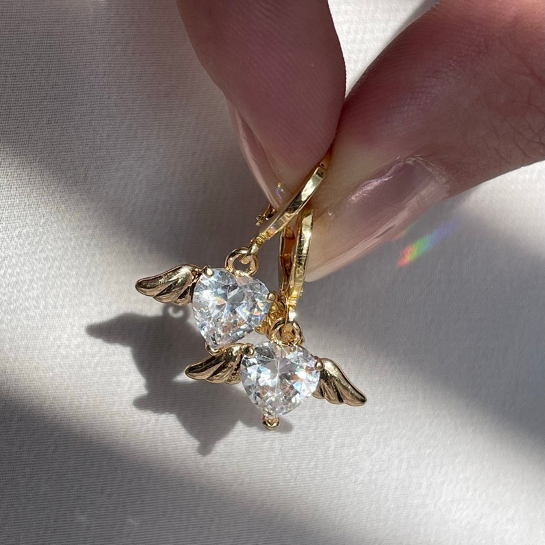 A close-up of a hand holding a pair of gold angel wings heart earrings, featuring sparkling cubic zirconia heart charms. The delicate wing details and radiant stones catch the sunlight, casting a subtle shadow on the white surface. These hypoallergenic earrings are perfect for everyday wear or as a meaningful gift for Christmas, birthdays, or special occasions. A beautiful choice for those who love celestial and angel-inspired jewelry.