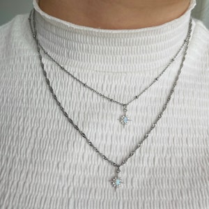 May include: Two silver necklaces with star-shaped pendants featuring a white opal stone. The necklaces are layered on a white shirt.