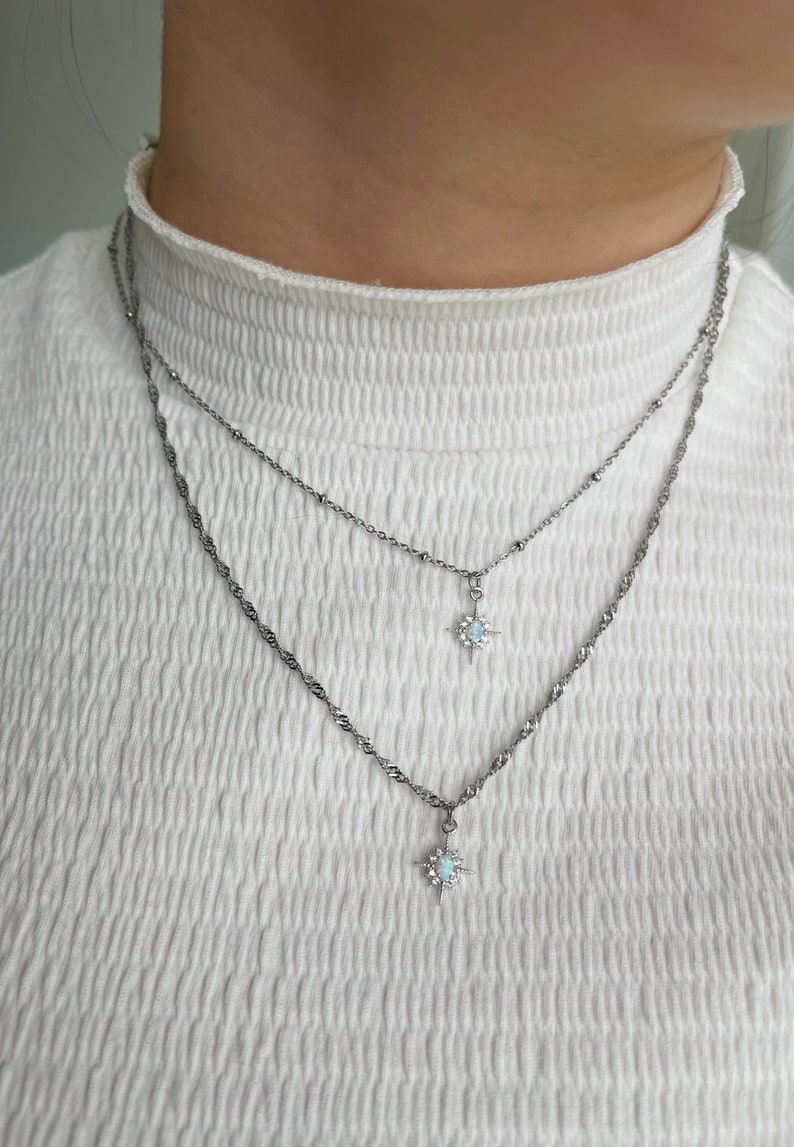 May include: Two silver necklaces with star-shaped pendants featuring a white opal stone. The necklaces are layered on a white shirt.