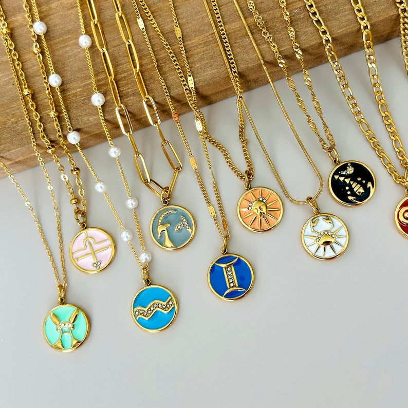 K&ouml;nnte beinhalten: Eine Auswahl goldfarbener Halsketten mit verschiedenen Anh&auml;ngerdesigns. Einige Anh&auml;nger zeigen Sternzeichen in Farben wie Rosa, Blau und Orange. Andere Halsketten haben Perlen oder einfache Kettenglieder. Die Halsketten werden auf einer hellen Oberfl&auml;che pr&auml;sentiert.