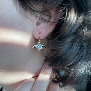 Może przedstawiać: Złote kolczyki koła z wiszącym sercem ozdobionym przezroczystymi kryształami i małymi skrzydłami.