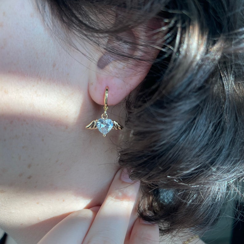 Może przedstawiać: Złote kolczyki koła z wiszącym sercem ozdobionym przezroczystymi kryształami i małymi skrzydłami.