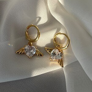 Może przedstawiać: Para złotych kolczyk&oacute;w koła z wiszącym sercem z kryształu i skrzydłami anioła.