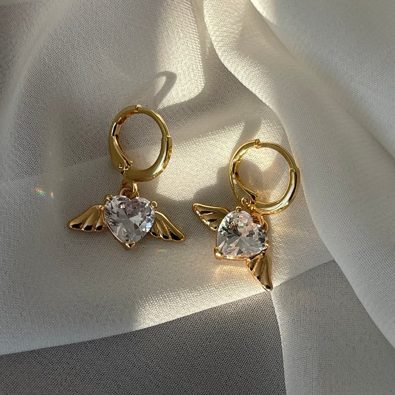 Może przedstawiać: Para złotych kolczyk&oacute;w koła z wiszącym sercem z kryształu i skrzydłami anioła.