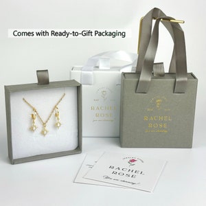 Może przedstawiać: Szare pudełko prezentowe z naszyjnikiem i kolczykami ze złota w środku. Pudełko jest oznaczone napisem "Rachel Rose" i ma mały wz&oacute;r r&oacute;ży. Pudełko stoi na białej powierzchni obok białej torby prezentowej z szarym uchwytem. Torba prezentowa jest r&oacute;wnież oznaczona napisem "Rachel Rose" i ma mały wz&oacute;r r&oacute;ży.