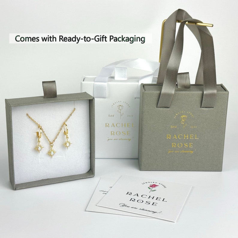 Może przedstawiać: Szare pudełko prezentowe z naszyjnikiem i kolczykami ze złota w środku. Pudełko jest oznaczone napisem "Rachel Rose" i ma mały wz&oacute;r r&oacute;ży. Pudełko stoi na białej powierzchni obok białej torby prezentowej z szarym uchwytem. Torba prezentowa jest r&oacute;wnież oznaczona napisem "Rachel Rose" i ma mały wz&oacute;r r&oacute;ży.