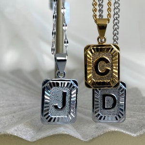 May include: Three rectangular pendant necklaces featuring the letters J, C, and D. The J and D pendants are silver-toned with a textured center and sunburst border. The C pendant is gold-toned with a matching textured design and border.