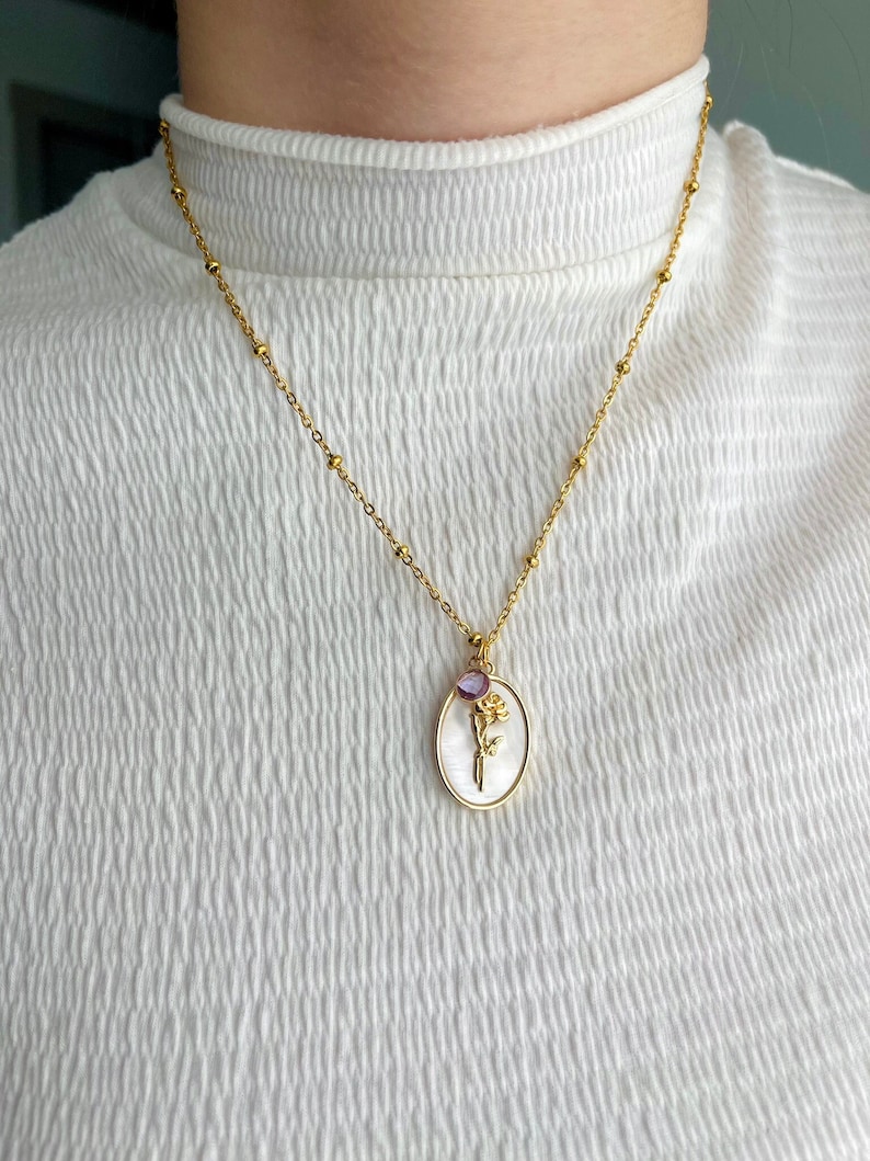 May include: A gold chain necklace featuring a satellite bead design and an oval pendant. The pendant has a white mother-of-pearl background with a gold-toned flower emblem and a small, round, purple gemstone set at the top of the oval frame.