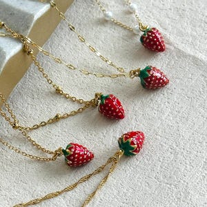 Peut inclure: Colliers dorés ornés de pendentifs fraises rouges. Les fraises ont des feuilles vertes et des détails de petites graines. Les chaînes sont de styles variés, avec des perles et des torsades. Présentés sur une surface texturée blanc cassé.