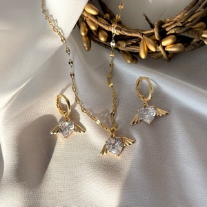 Może przedstawiać: Zestaw złotego naszyjnika i kolczyk&oacute;w z serduszkowym wisiorem i pasującymi kolczykami. Wisiorek i kolczyki mają przezroczysty kryształ z małymi skrzydełkami po obu stronach.