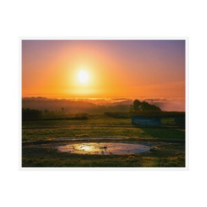 Puede incluir: Una salida del sol sobre un campo con una estructura de piedra circular en primer plano. El sol es de color naranja brillante y el cielo es un degradado de naranja, rosa y morado.