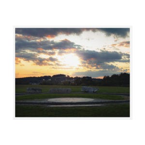 Puede incluir: Una vista panorámica de un campo de hierba con una estructura circular de piedra en el centro. El cielo es una mezcla vibrante de rosa, naranja y azul con nubes esponjosas. El sol se pone detrás de una línea de árboles en la distancia.