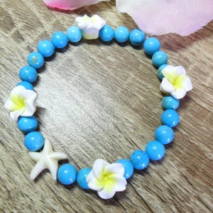 Puede incluir: Una pulsera de cuentas turquesas con detalles de estrellas de mar y flores blancas. La pulsera presenta cuentas redondas de color azul claro y abalorios blancos de flores y estrellas de mar. Joyería con temática veraniega.