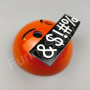 May include: An orange emoji with a black bar across it, displaying white symbols. The emoji has a curved mouth and a small, slanted eye. The black bar has the symbols: $!#%&.