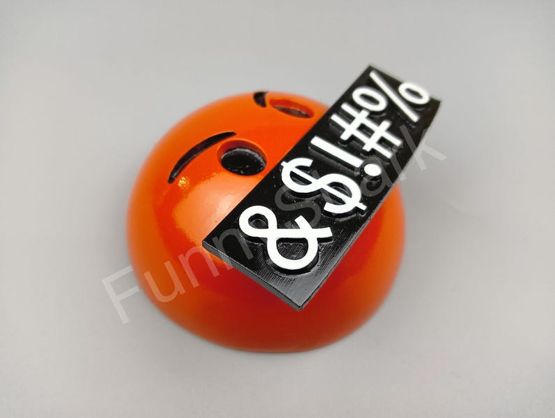 May include: An orange emoji with a black bar across it, displaying white symbols. The emoji has a curved mouth and a small, slanted eye. The black bar has the symbols: $!#%&.
