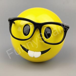 Puede incluir: Hucha amarilla con cara sonriente, gafas de pasta negra y una amplia sonrisa.