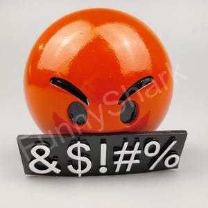 May include: A shiny orange emoji-style sphere with black eyes and eyebrows, and a black rectangular base with white symbols: & $ ! # %. The emoji has an angry expression.