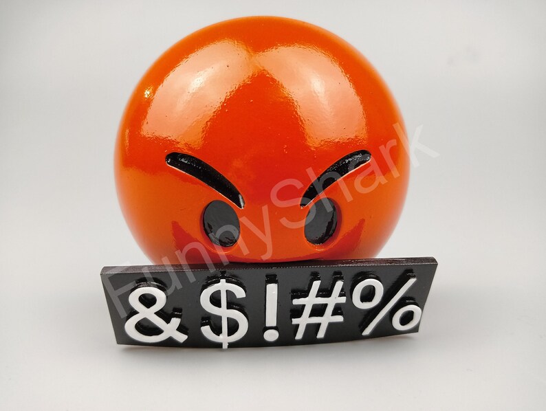 May include: A shiny orange emoji-style sphere with black eyes and eyebrows, and a black rectangular base with white symbols: & $ ! # %. The emoji has an angry expression.