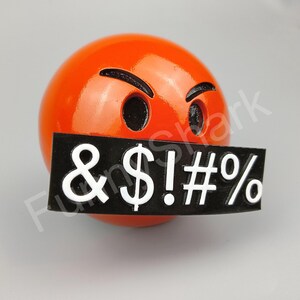 May include: A spherical orange object with a black sign covering its mouth. The sign displays the symbols & $ ! # % in white. The object has black eyebrows and eye holes, giving it an angry expression.