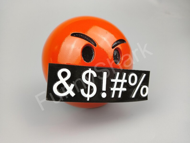 May include: A spherical orange object with a black sign covering its mouth. The sign displays the symbols & $ ! # % in white. The object has black eyebrows and eye holes, giving it an angry expression.